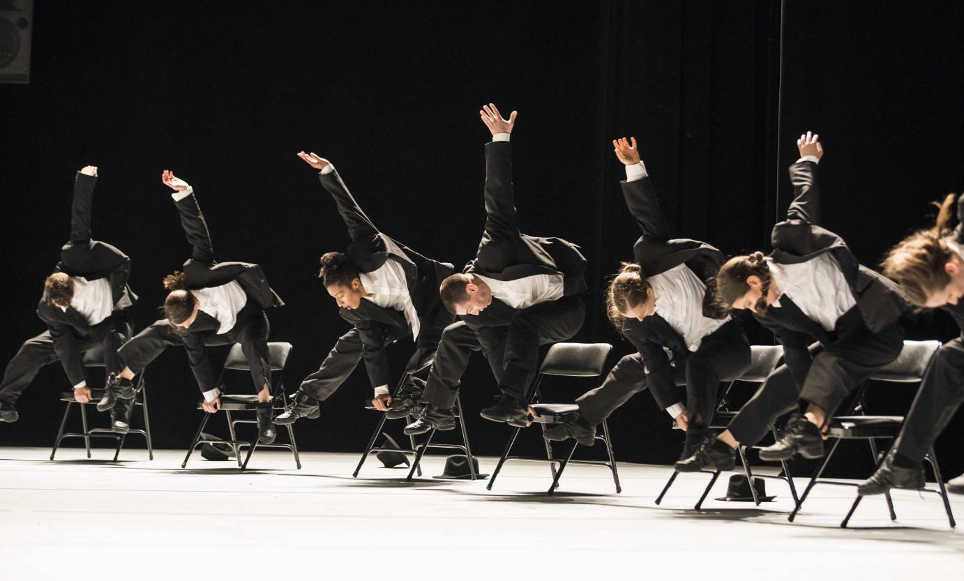 Sixteen dancers in black suits lift themselves off black folding chairs